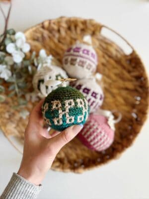 A hand holds a crocheted green ornament over a woven basket filled with crochet tree decorations in various colors.