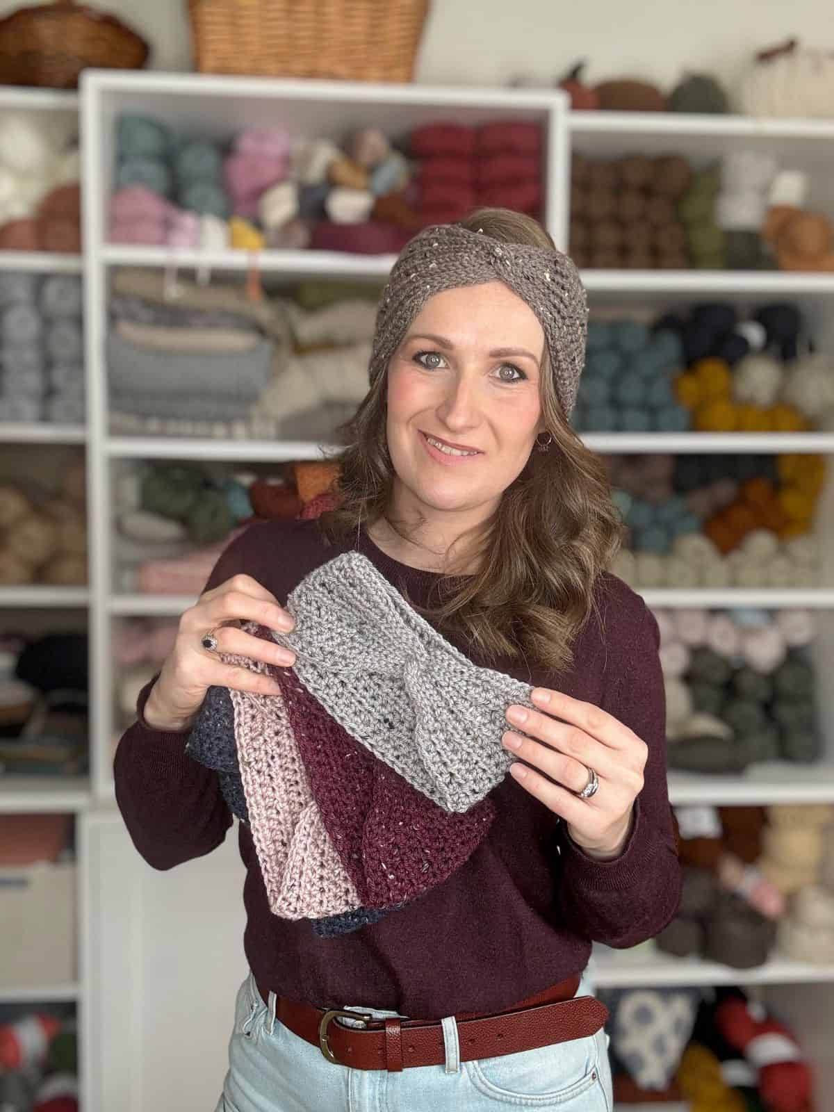 A woman in a maroon sweater holds crocheted headbands, showcasing her easy crochet ear warmer pattern, in front of shelves filled with yarn and other crocheted items.