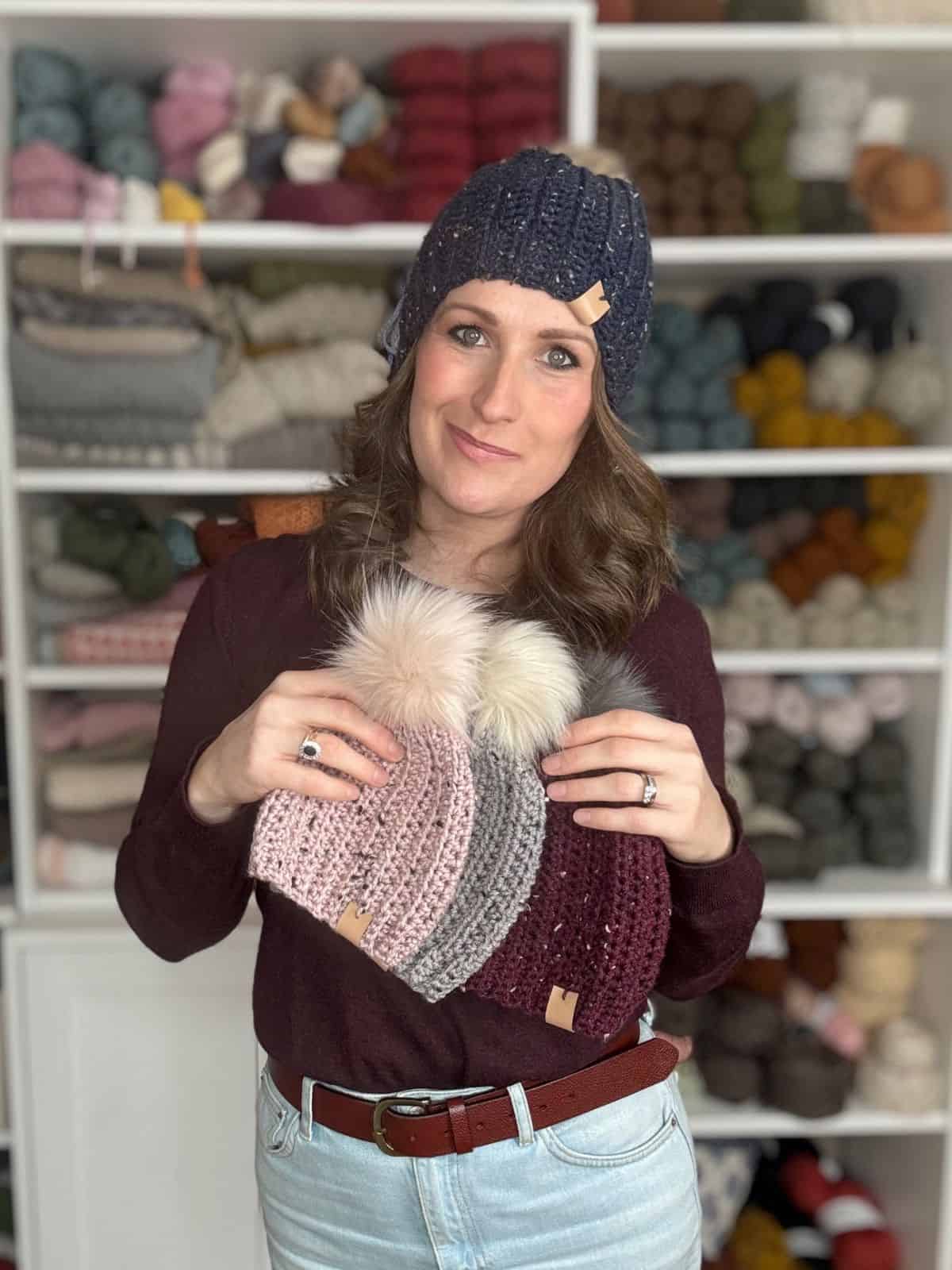 A person in a crochet hat, made using an easy crochet hat pattern, holds three beanies with pom-poms. Shelves with assorted yarns are in the background.