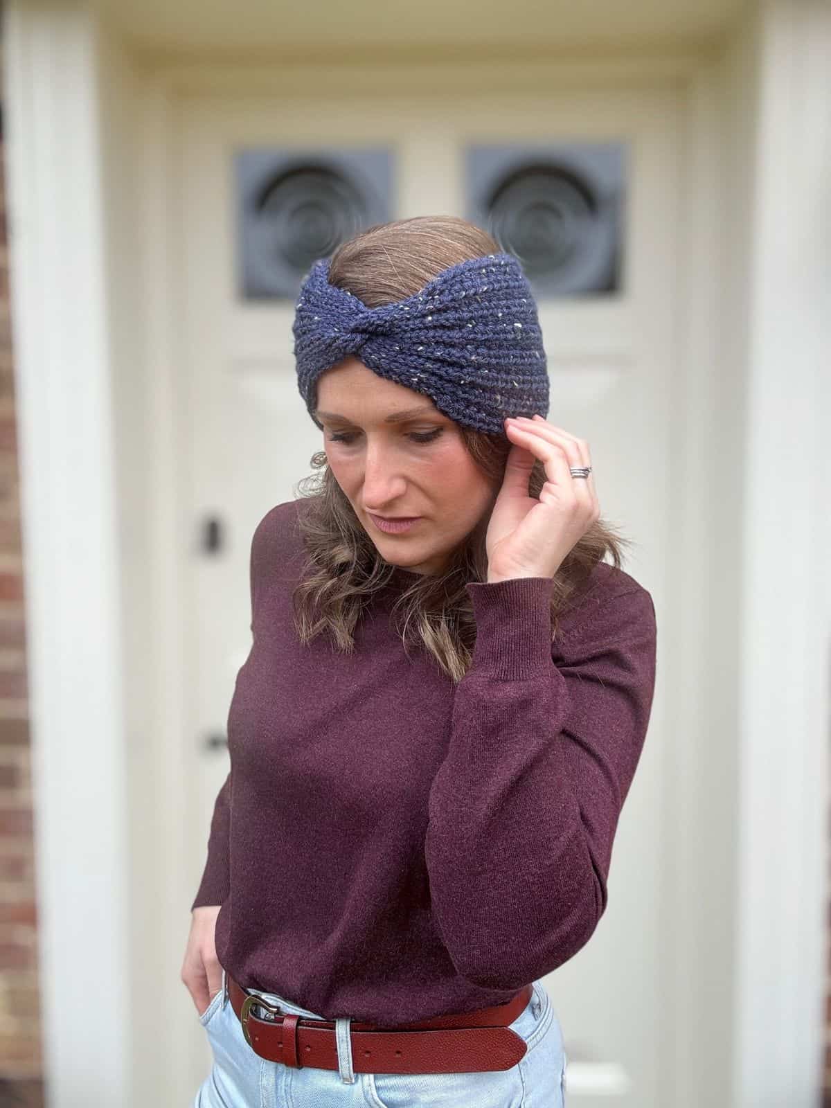 A woman wearing a crochet headband and maroon sweater poses gracefully in front of a white door, showcasing an easy crochet ear warmer pattern that complements her cozy style.