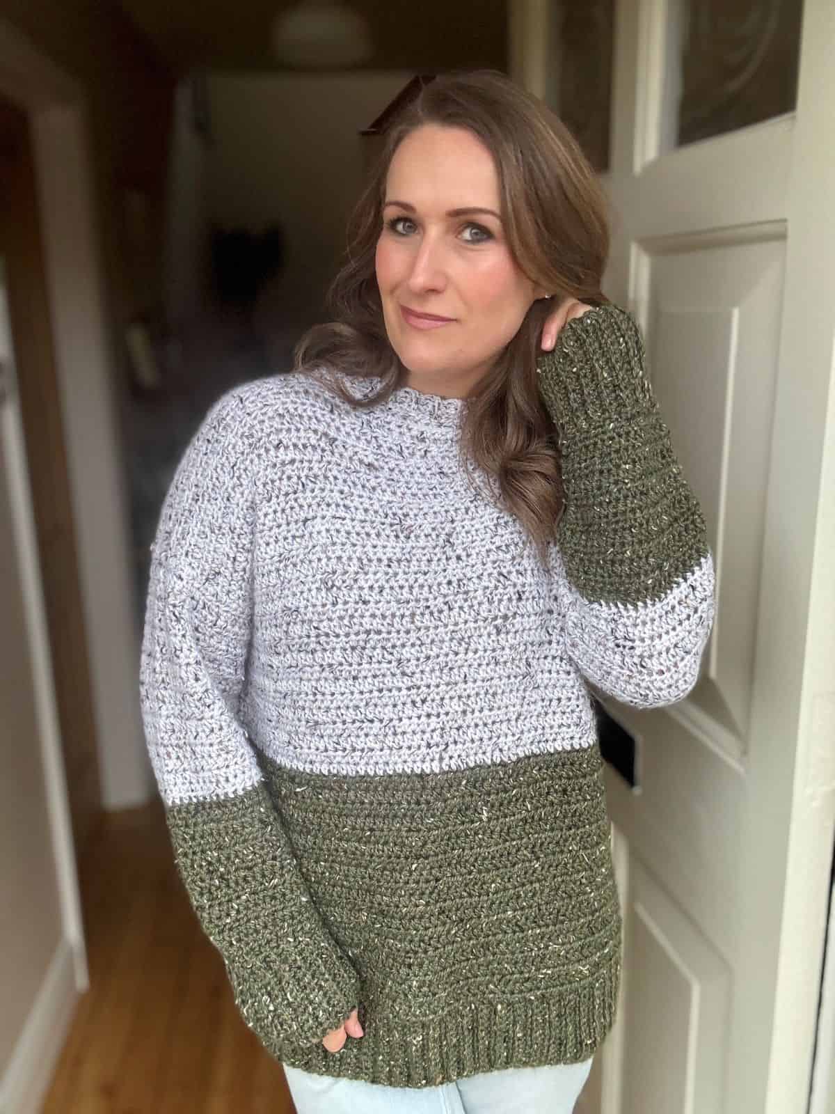 A woman stands indoors wearing an oversized crochet sweater with mixed green and gray colors, posing with her hand on her neck. She has long, wavy hair and is looking at the camera.