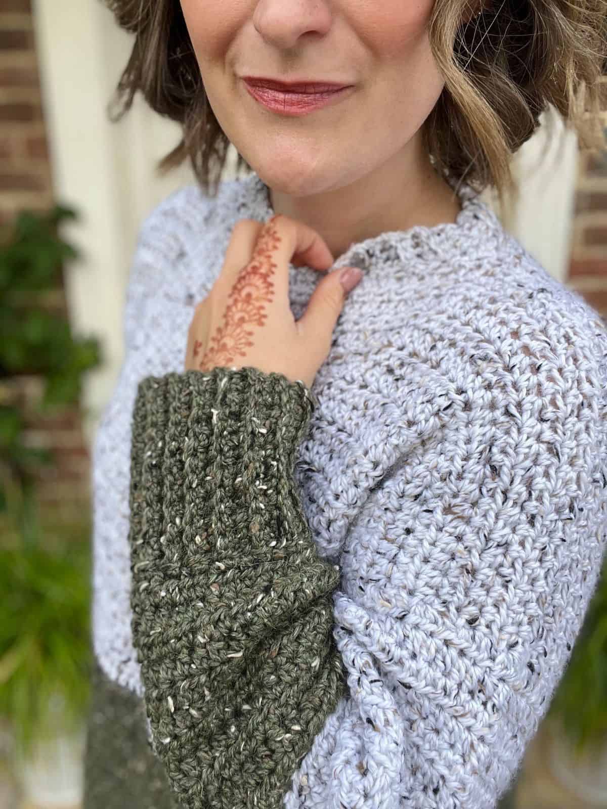 A person wearing an oversized, multicolored crochet sweater with a crochet pattern displays intricate henna on their hand.