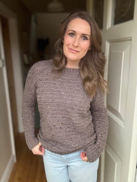 Person with long hair wearing a crocheted sweater and jeans, standing indoors by a door, smiling slightly.