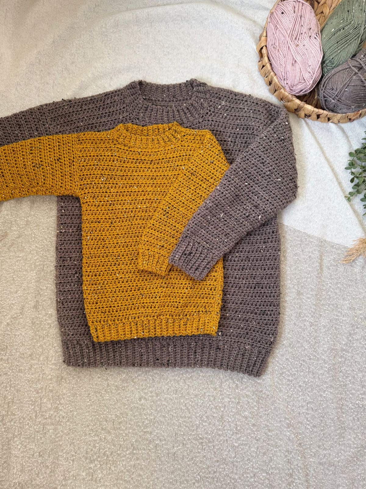 Two crocheted sweaters lie on a beige surface: a mustard sweater on top of an oversized gray one. Nearby, a basket filled with green, pink, and brown yarn hints at the variety of crochet patterns used.