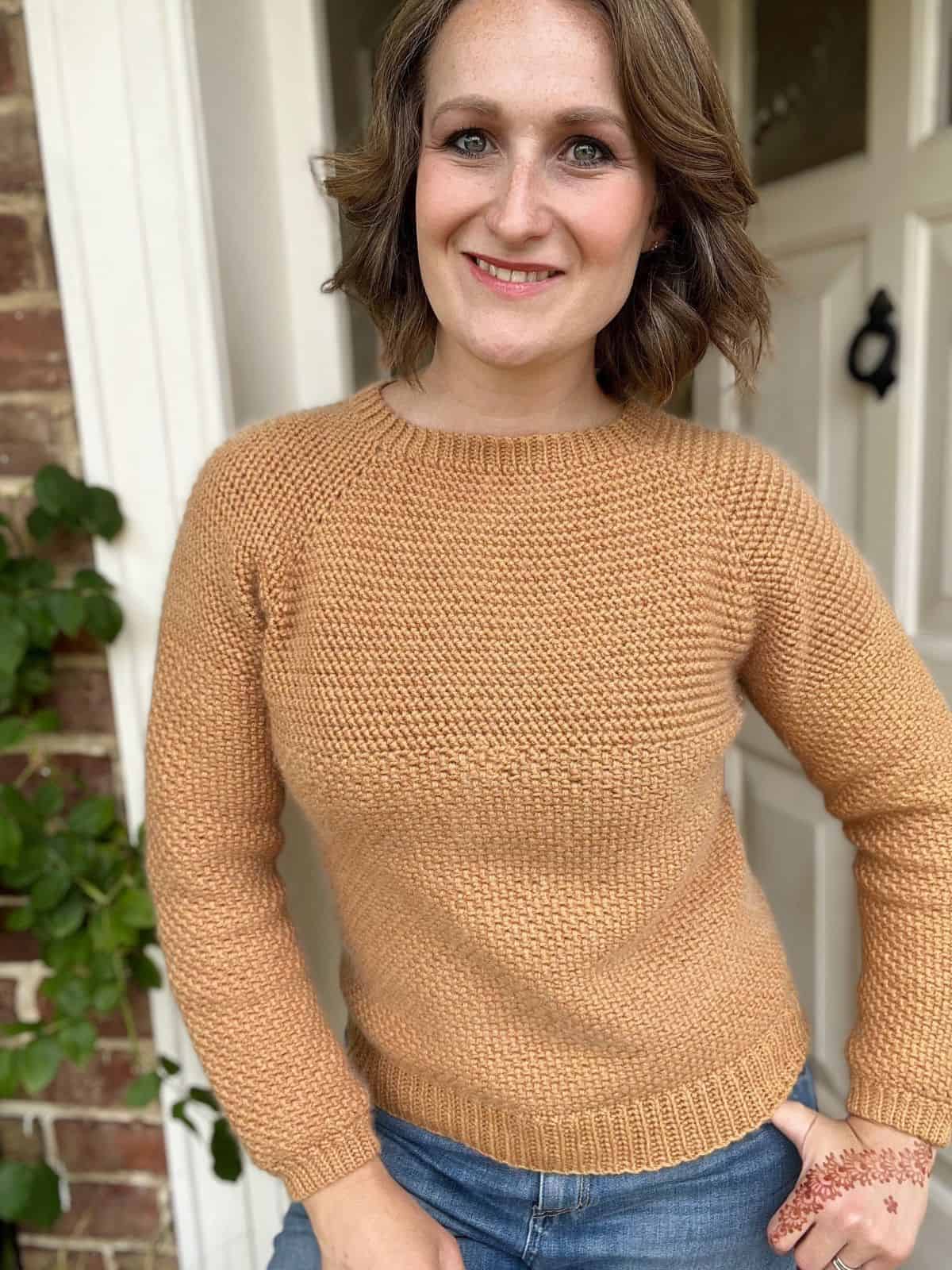 A person smiling, adorned in a crocheted beige sweater and jeans, stands in front of a white door with plants on the side. The cozy look could easily inspire anyone seeking a men's crochet sweater pattern.