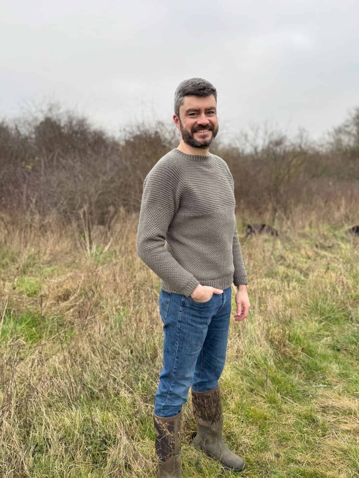 A man stands in a grassy field, wearing a stylish grey crochet sweater, blue jeans, and brown boots, with trees gracefully framing the background.