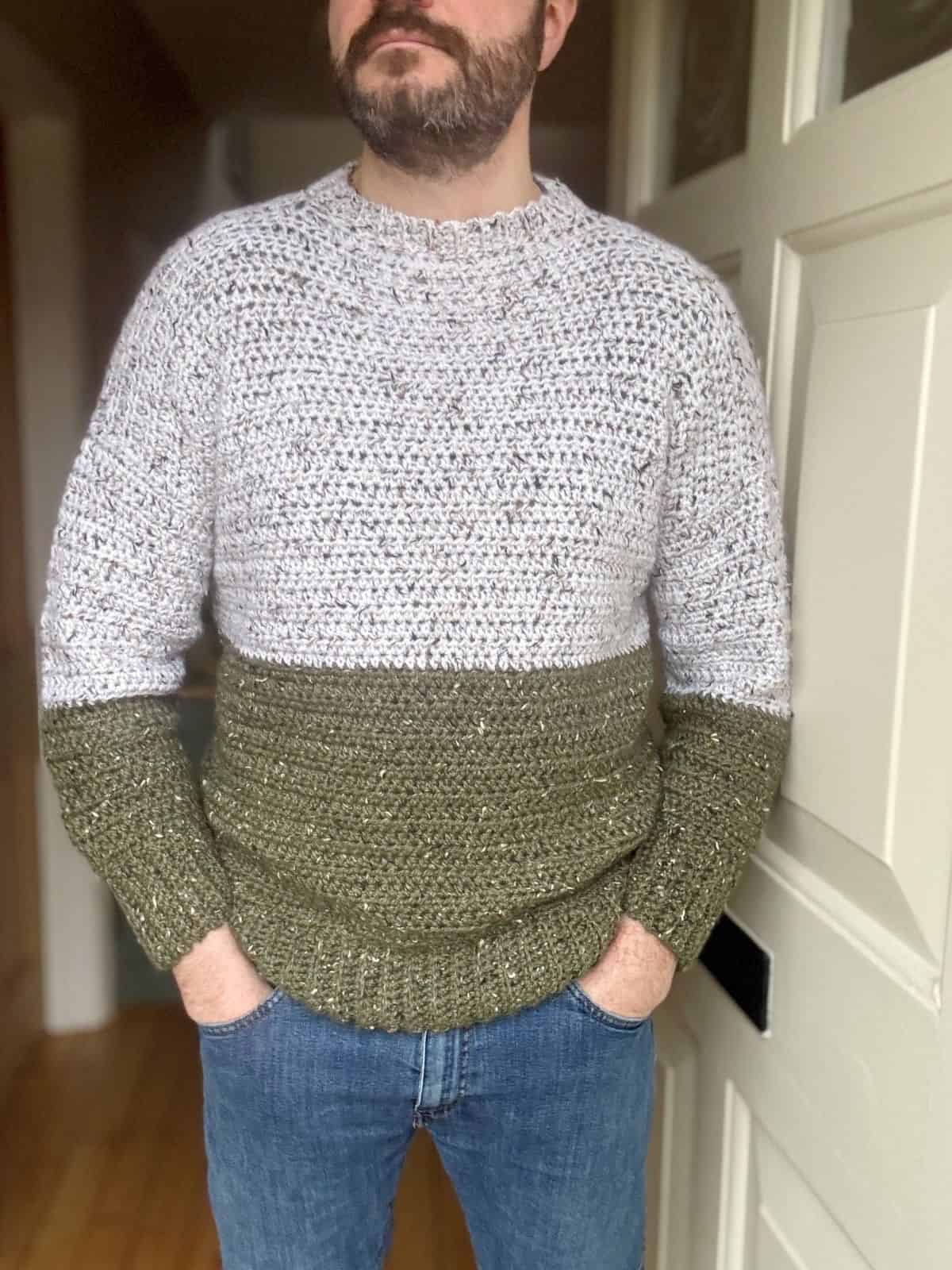 A man stands with hands in pockets, donning an oversized crochet sweater with white and green sections. The crochet pattern adds a unique texture, complementing the cream-colored door behind him.