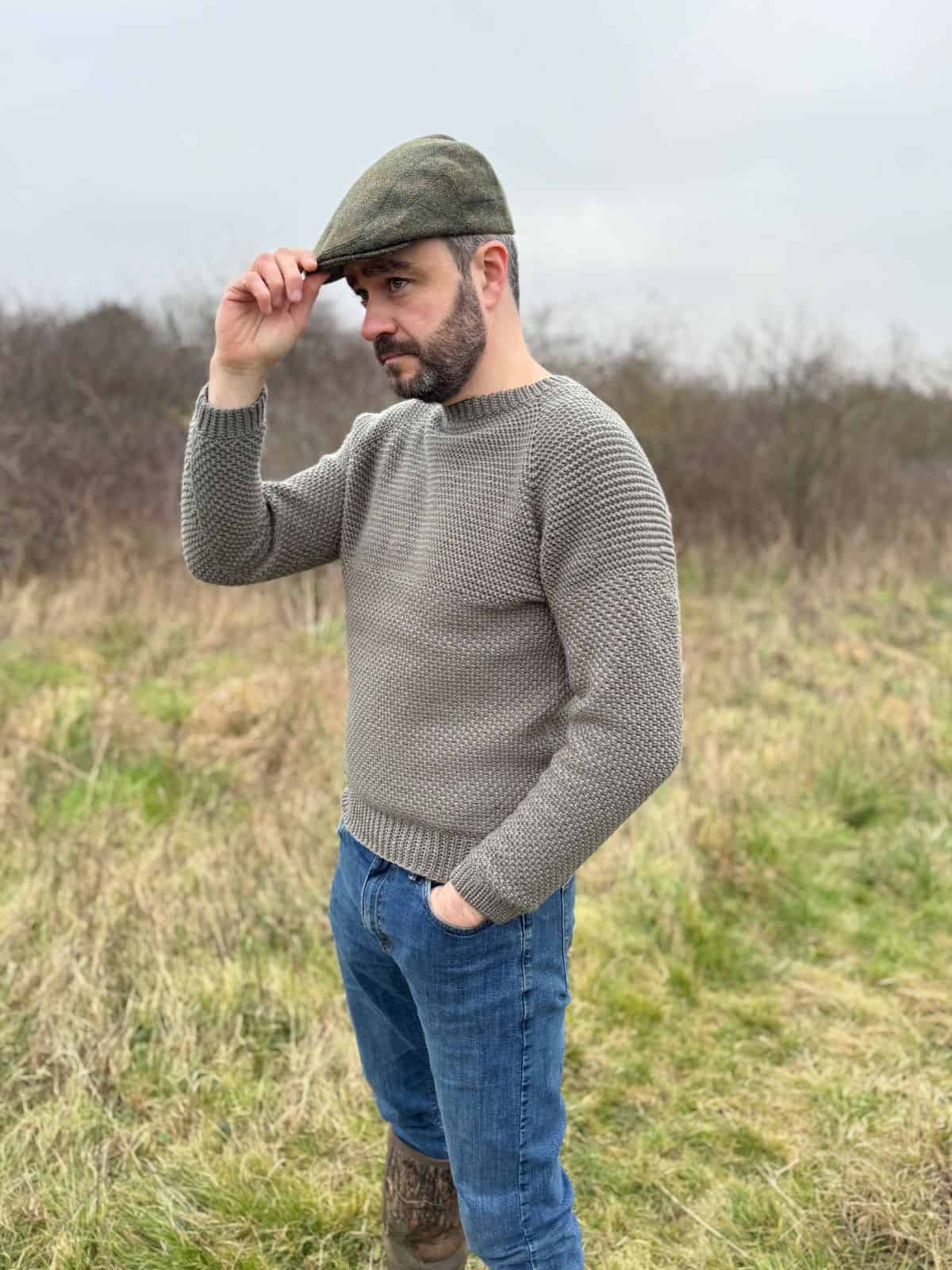 A man in a grey crochet sweater, jeans, and flat cap stands in a grassy field, adjusting his cap. The men's sweater pattern complements the overcast sky in the background.