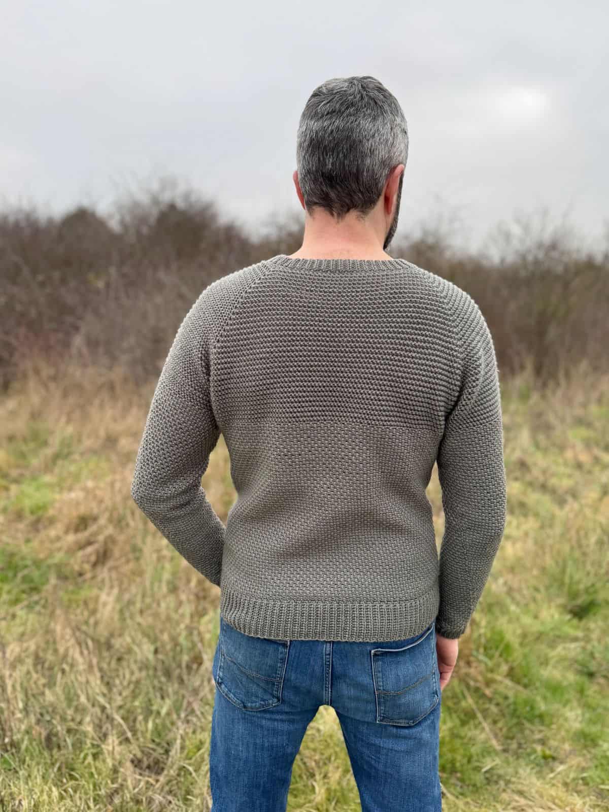 A person with gray hair, wearing a blue jeans and a textured gray crochet sweater featuring a men's crochet pattern, stands with their back to the camera in a grassy field under overcast skies.