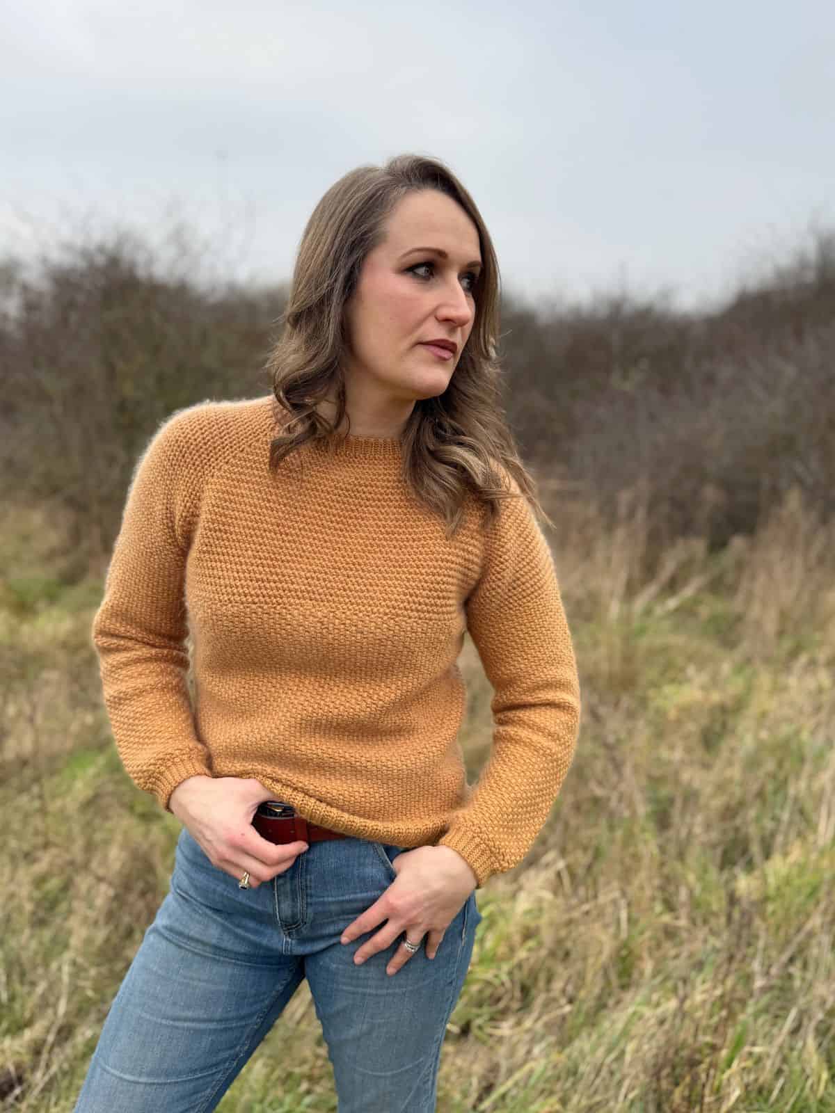 A person stands in a field wearing a mustard men's crochet sweater pattern and blue jeans, looking to the side. The background is grassy with shrubs.