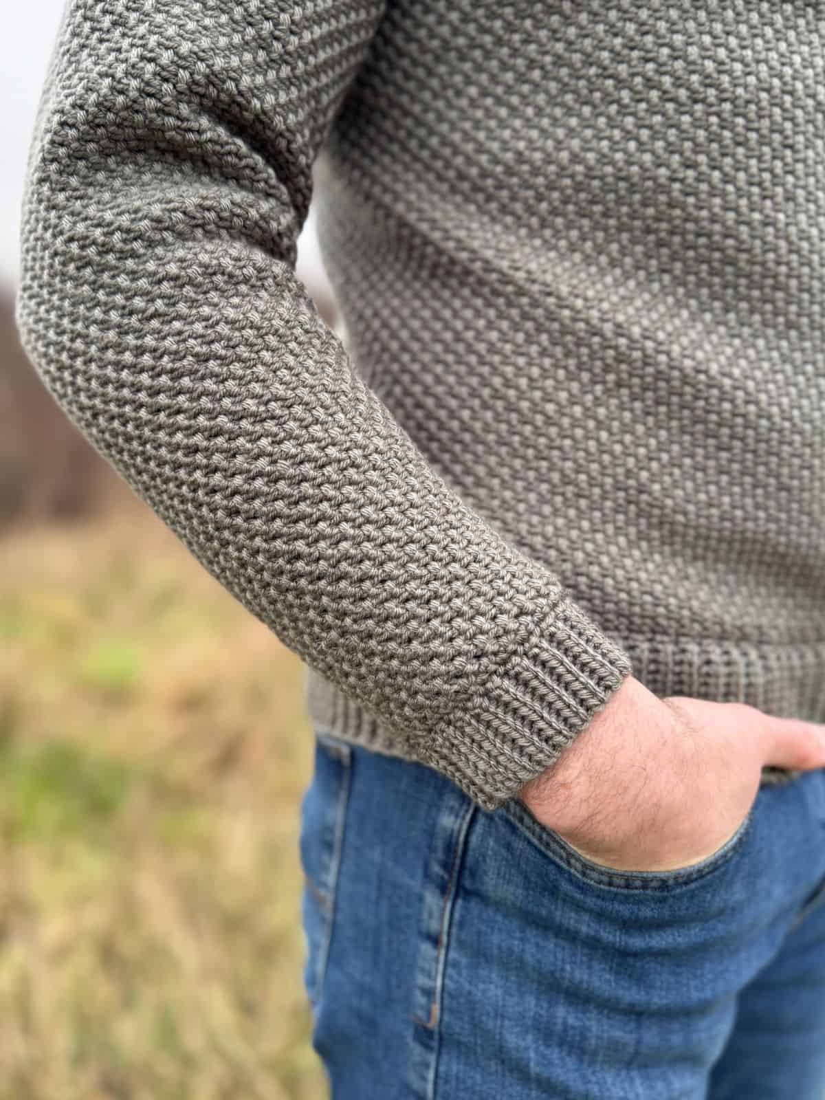 A person is casually standing in a grassy area, dressed in blue jeans and a textured gray crochet sweater that follows a men's pattern. One hand rests comfortably in their pocket.