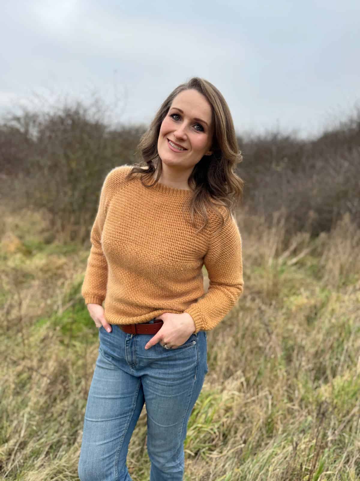 A person wearing a tan crochet sweater pattern and jeans stands smiling in a grassy, bushy outdoor area.