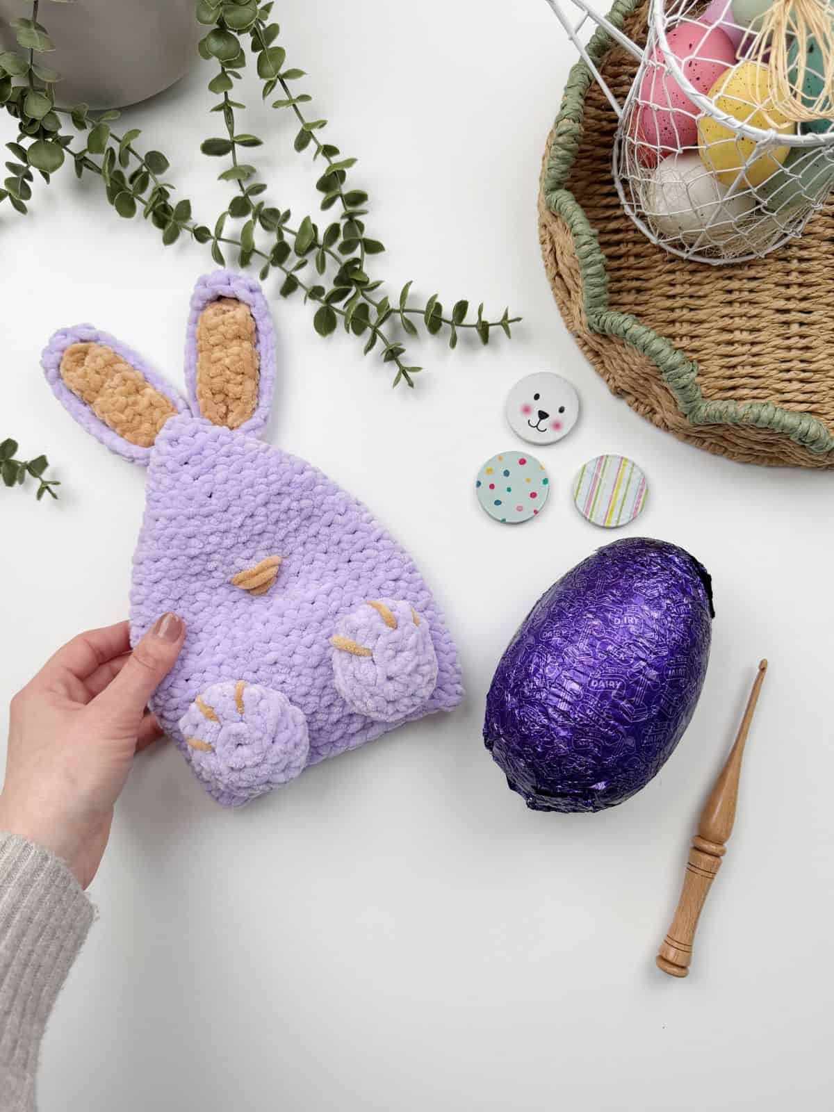 A hand holds a crocheted purple bunny pouch crafted from a crochet bunny pattern beside a purple foil-wrapped egg, two buttons, a crochet hook, and a basket of colorful eggs on a white surface with greenery.