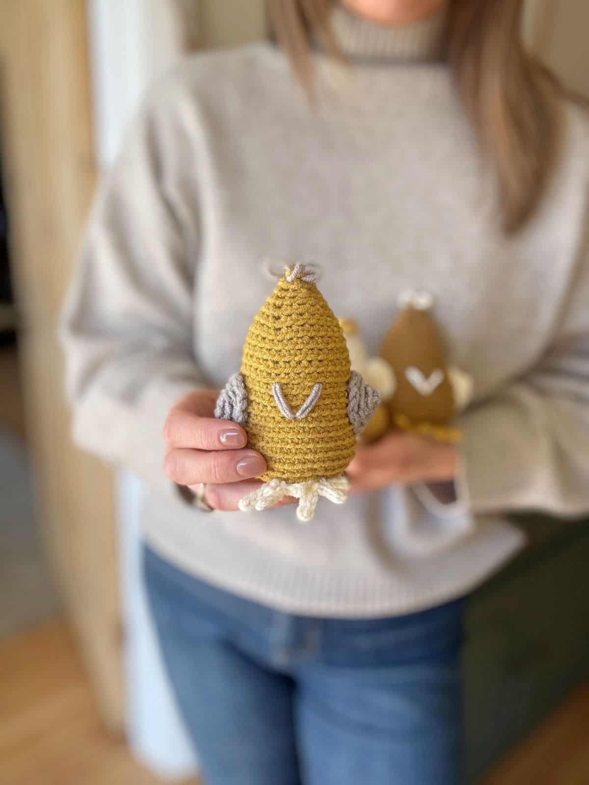 A person is holding a small, crochet chickens, created from a crochet chicken pattern. They are dressed in a beige sweater and jeans. In the background, another similar creation peeks into view.