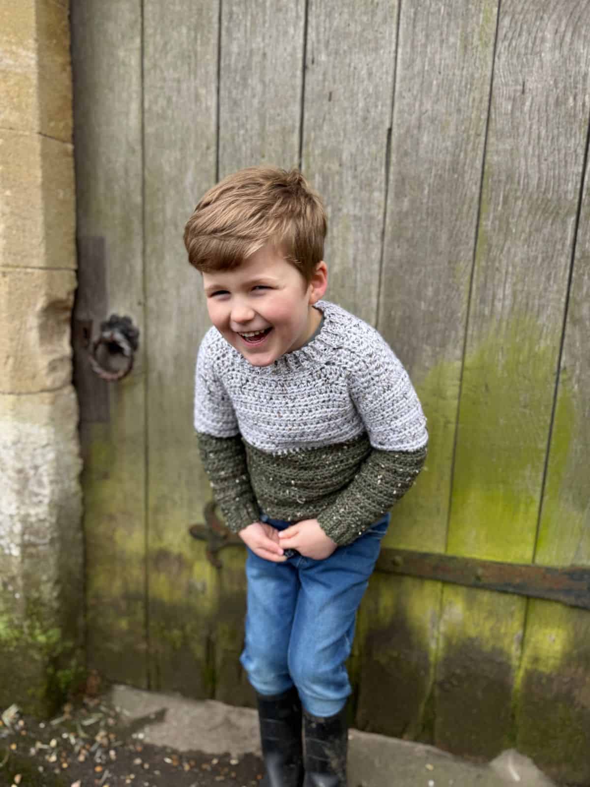 A child with short hair is laughing while standing in front of a weathered wooden door. They are wearing the easiest sweater to crochet, featuring gray and green hues, blue jeans, and black boots.