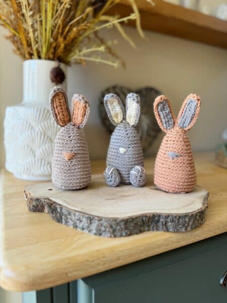 Three exquisite crochet bunny patterns are showcased on a wooden slice. With elegant gray and pink hues, they sit gracefully on a wooden table adorned with decorative items in the background.