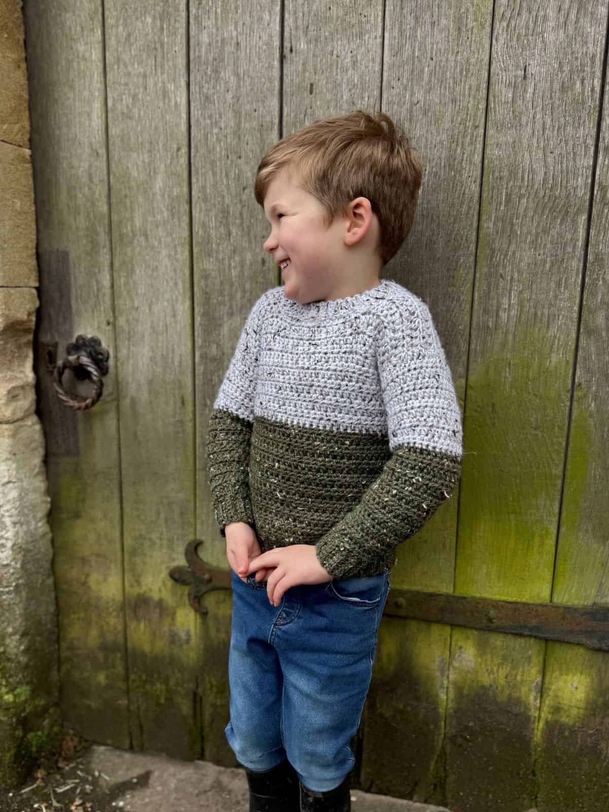 A child in a cozy gray and green crochet sweater, which looks like the easiest sweater to crochet, stands smiling beside a wooden door, gazing thoughtfully to the side.