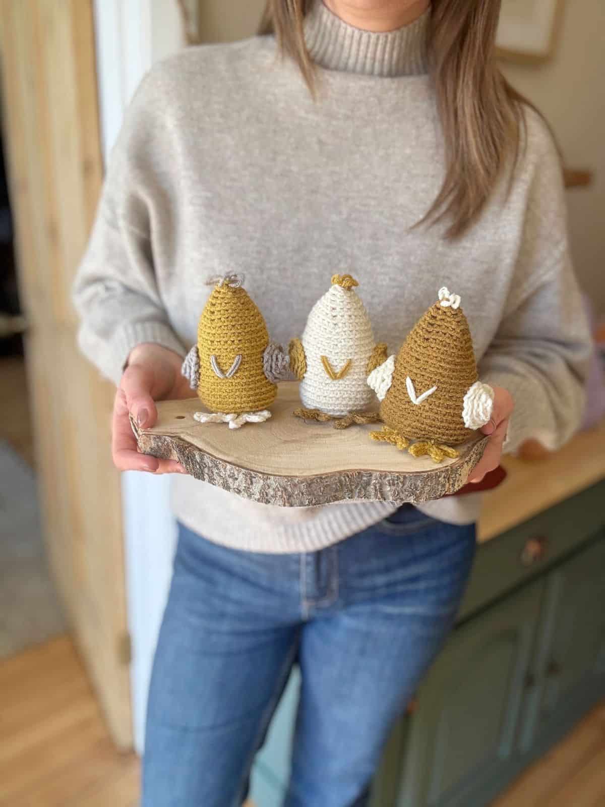 A person holds a wooden tray showcasing three delightful crochet chicken ornaments, intricately made in gray and brown hues. The craftsmanship hints at a crochet chicken pattern, adding a cozy charm to the scene.