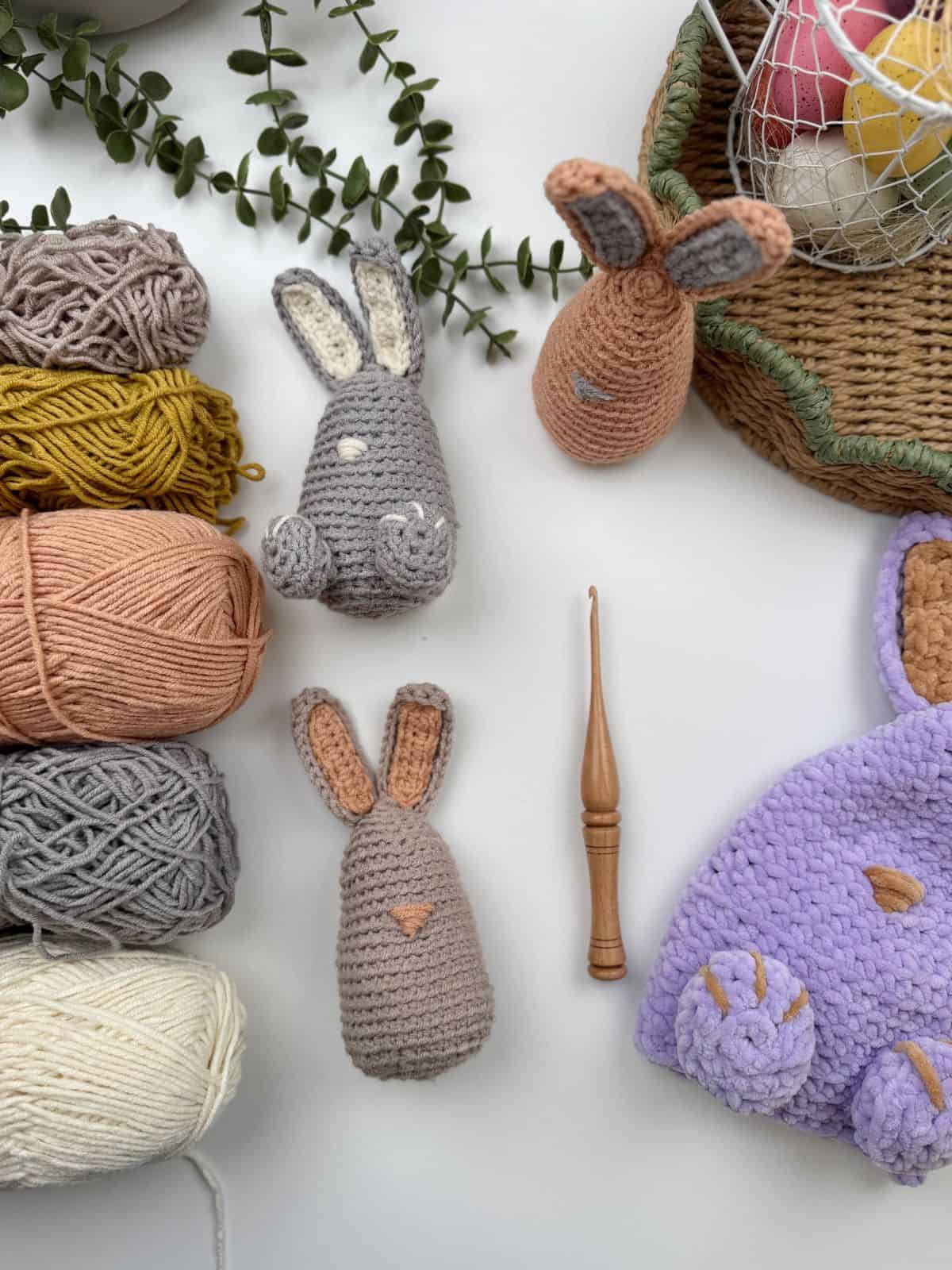 Crocheted rabbit figures crafted from a delightful crochet bunny pattern sit alongside a wooden crochet hook on a white surface, surrounded by skeins of yarn in various colors. A basket with colored eggs and leaves is visible nearby.