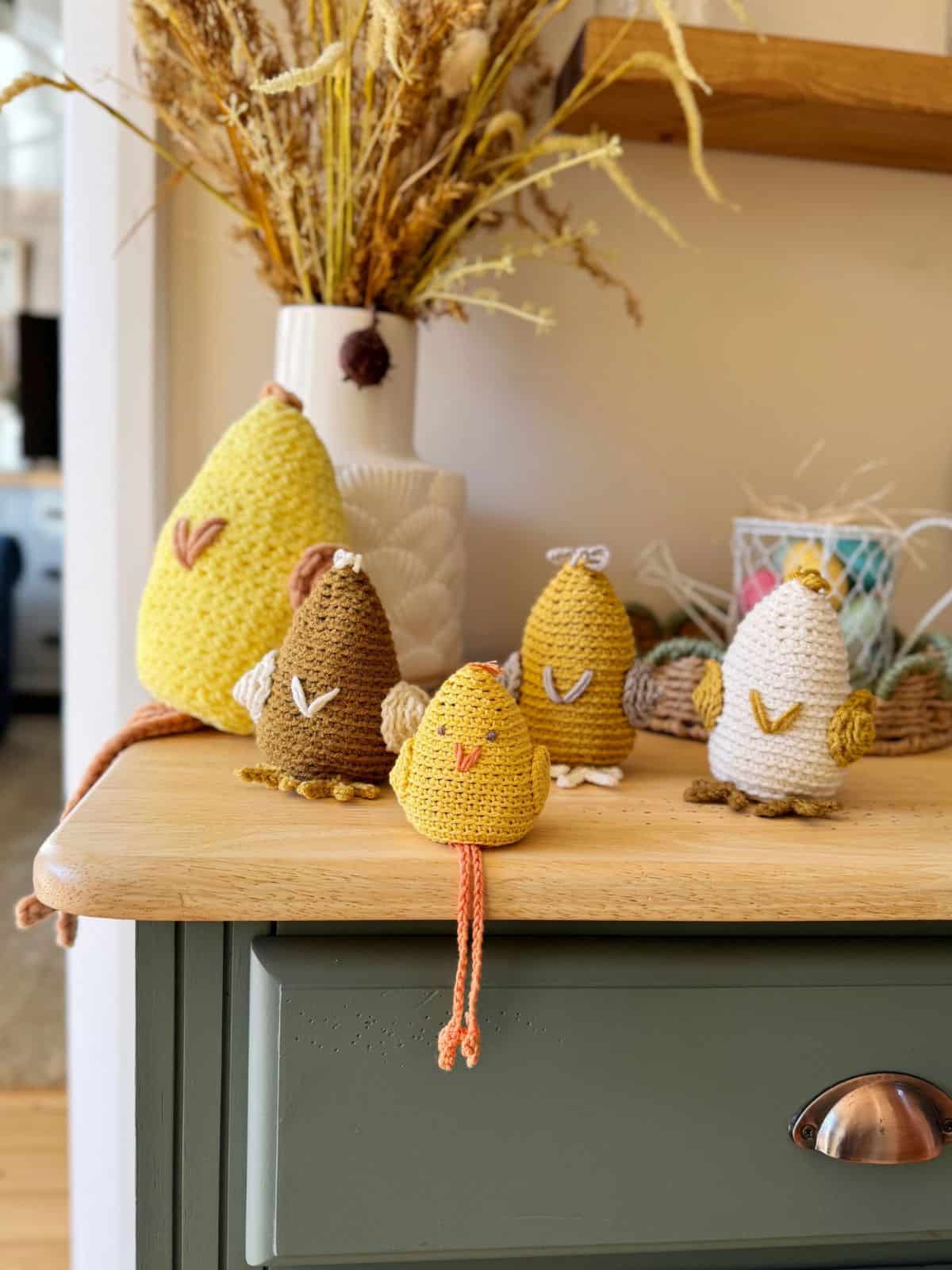 Crocheted birds in various colors, crafted with a delightful crochet chicken pattern, sit on a light wooden table. A vase with dried plants is in the background.