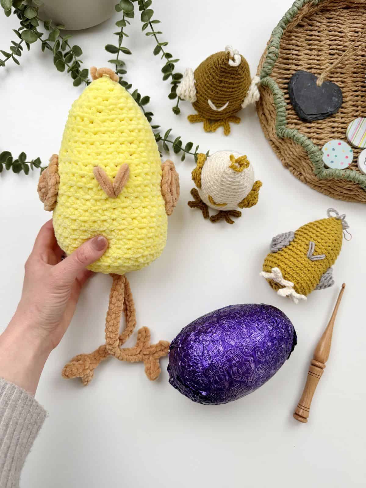 A hand holds a yellow crocheted bird crafted from a crochet chicken pattern beside a purple foil-wrapped chocolate egg, two smaller crocheted birds, and a basket adorned with a chalkboard heart and buttons.