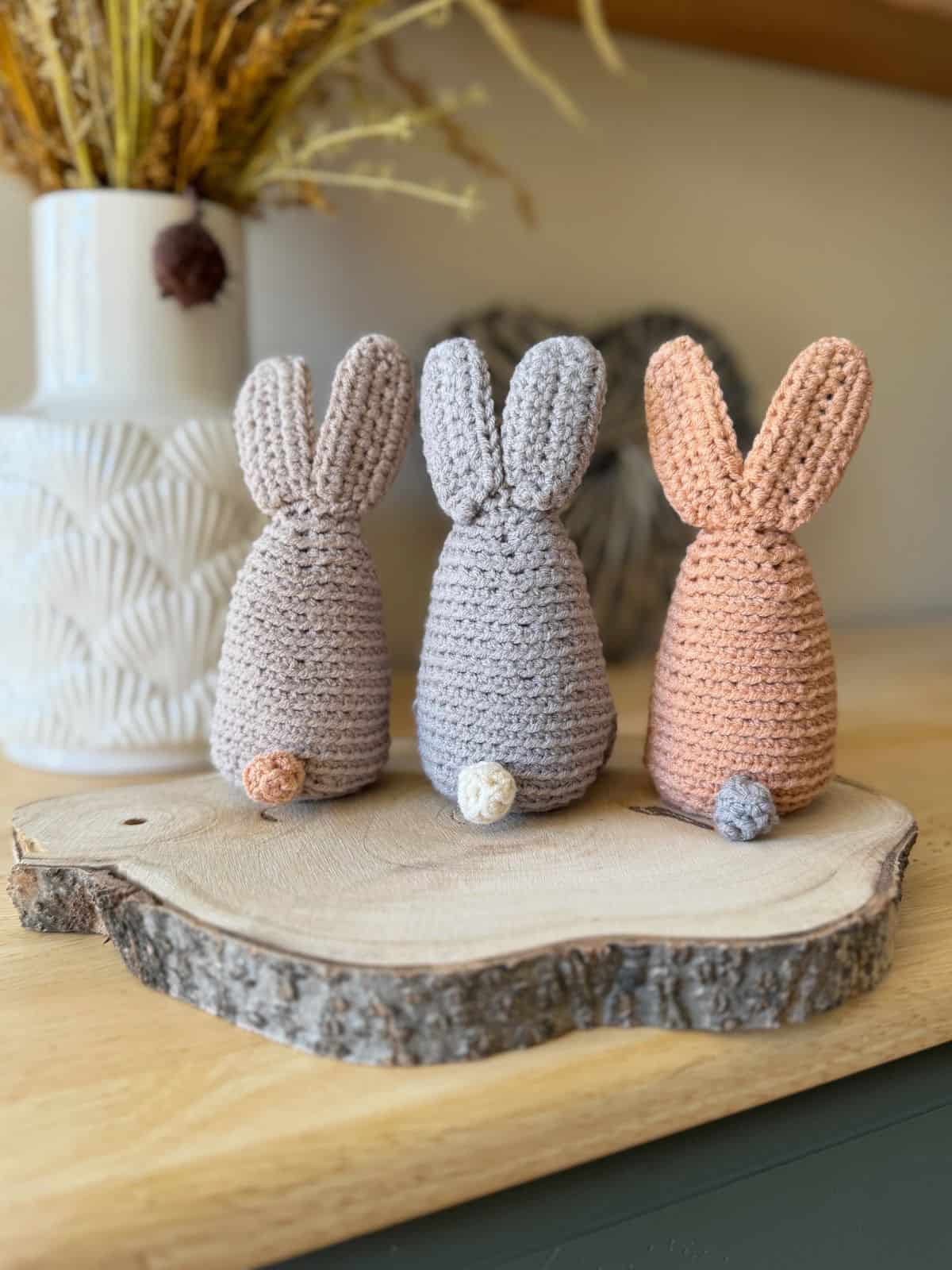 Three crocheted bunnies in gray and beige, crafted using a delightful crochet bunny pattern, sit on a wooden slab. They are displayed on a light wood surface with a decorative vase in the background.