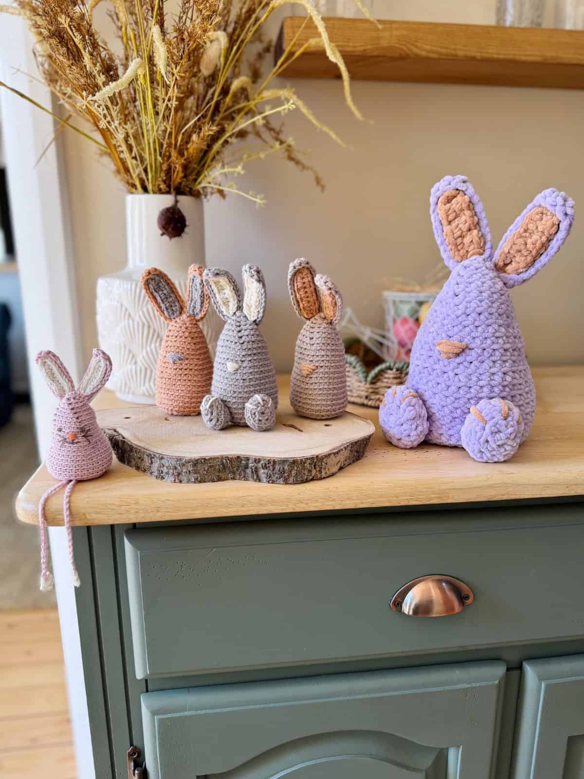 Five crocheted bunnies in various sizes and colors are displayed on a wooden surface, showcasing their unique charm alongside a vase of dried flowers. Their intricate designs could easily be inspired by a delightful crochet bunny pattern.