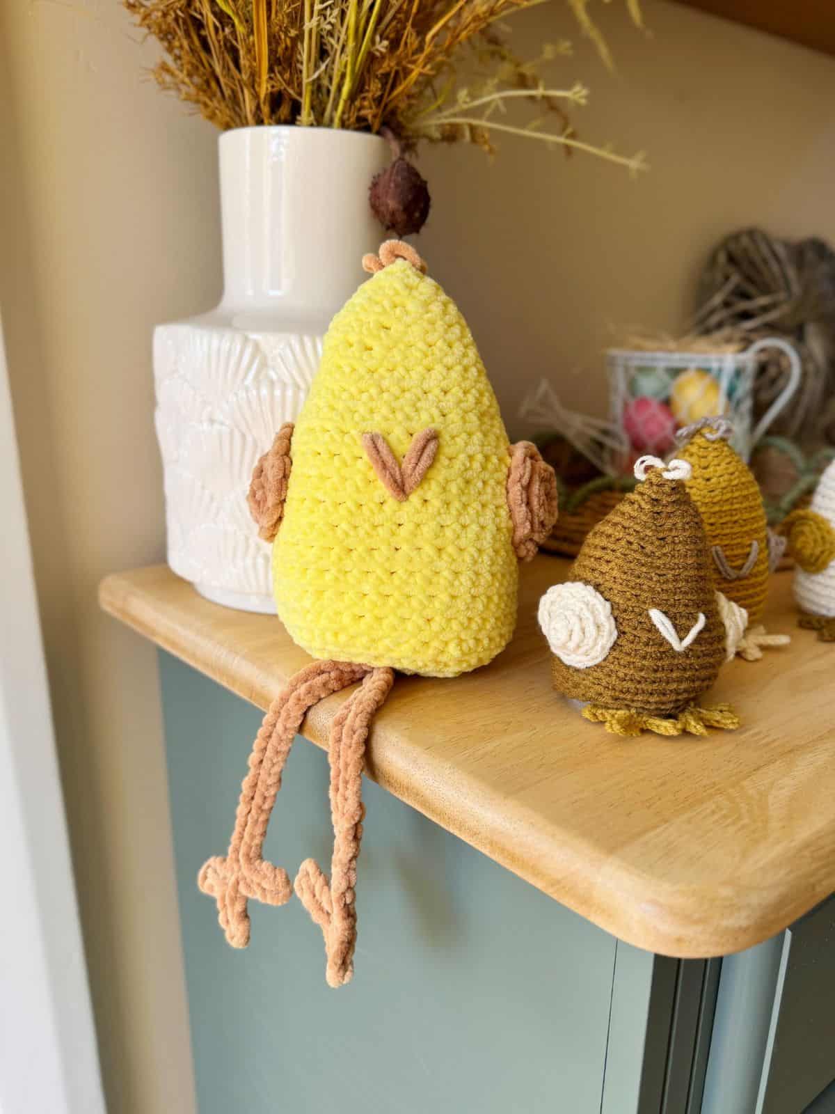 A yellow knitted bird with brown feet and wings, crafted using a crochet chicken pattern, sits on a shelf next to a vase and smaller knitted birds.