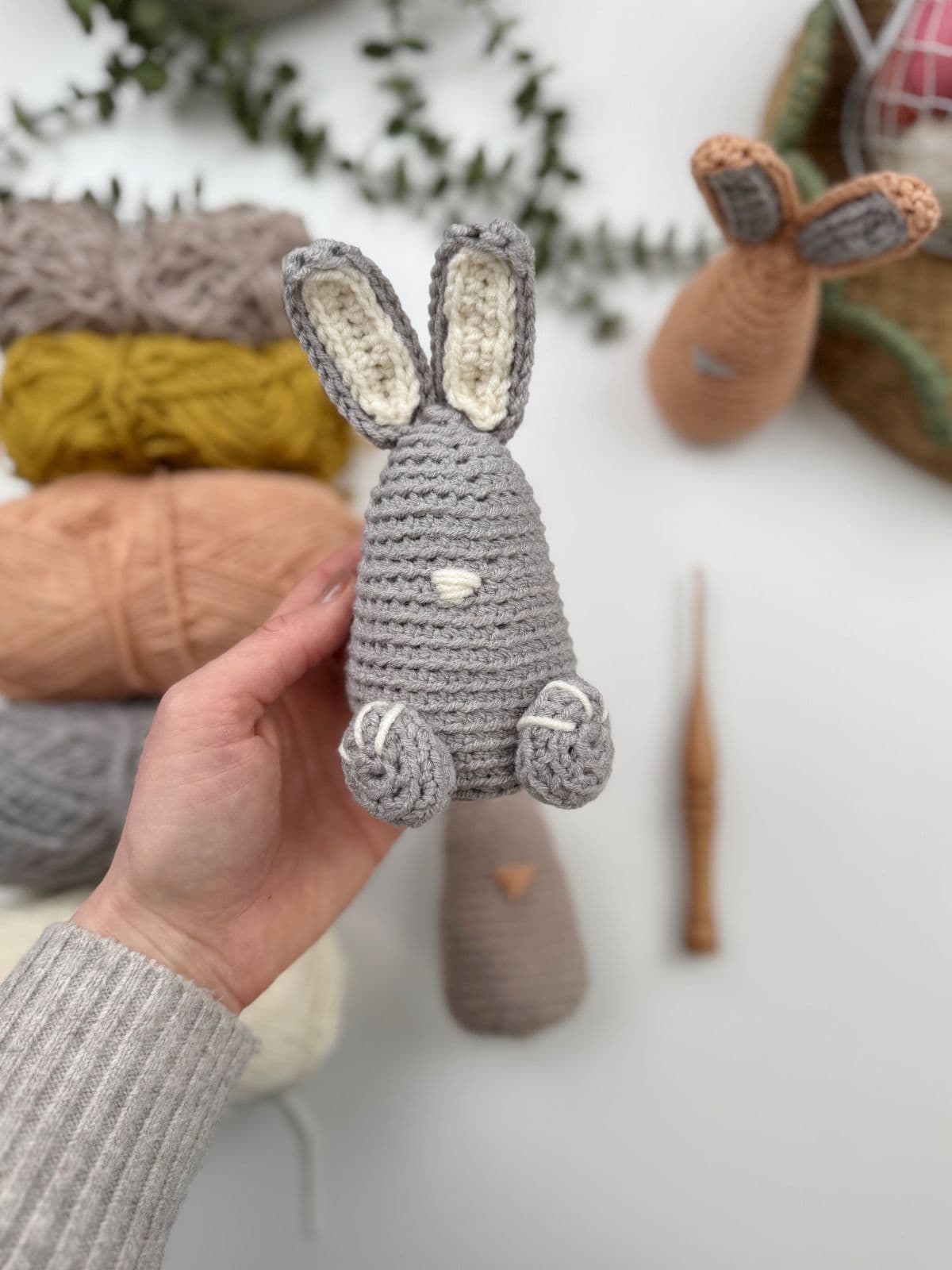 A hand holds a crocheted gray bunny with white details, surrounded by yarn balls and a crochet hook on a white surface—a perfect example of the crochet bunny pattern in action.