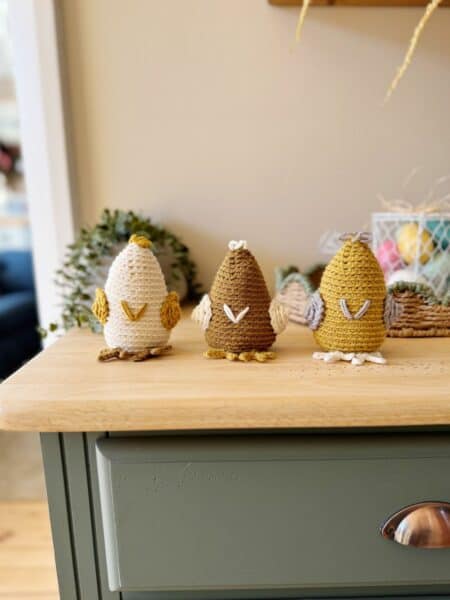 Three crocheted chicken figures in white, yellow, and brown are lined up on a wooden surface with decorative items in the background.