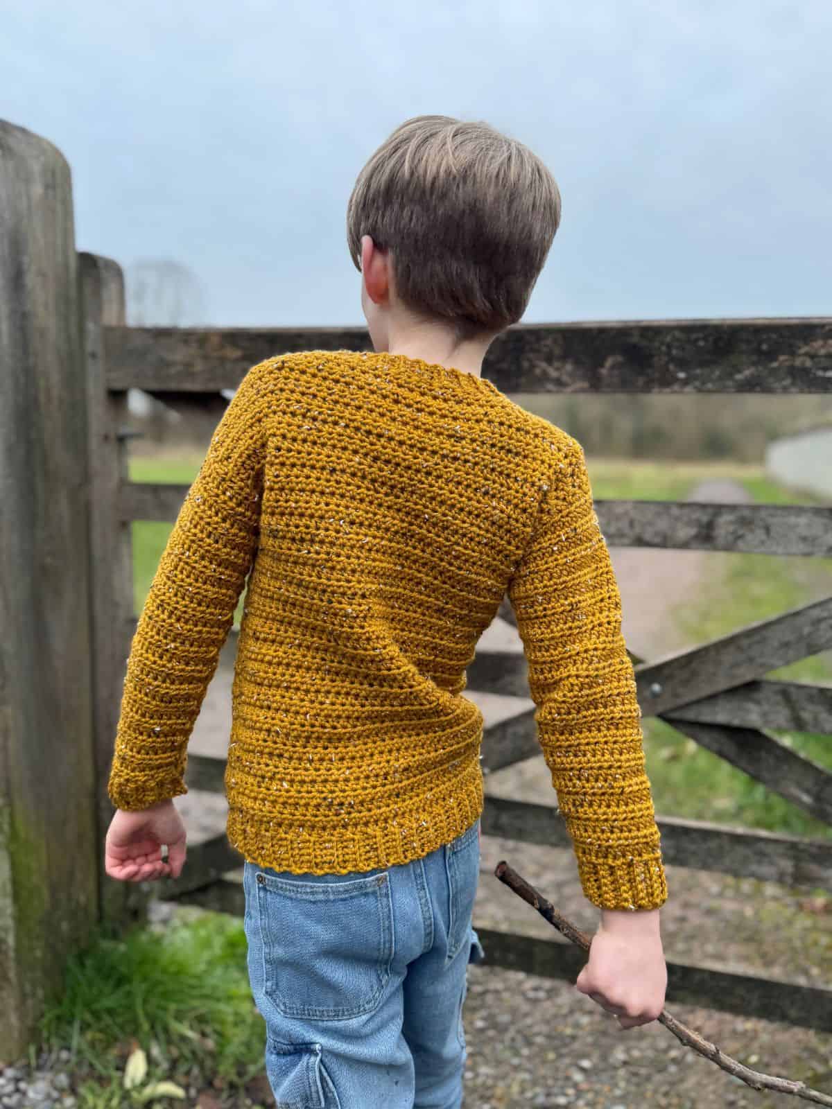 A child wearing a mustard-colored crocheted sweater, perhaps the easiest sweater to crochet, and jeans stands holding a stick in front of a wooden gate on a cloudy day.