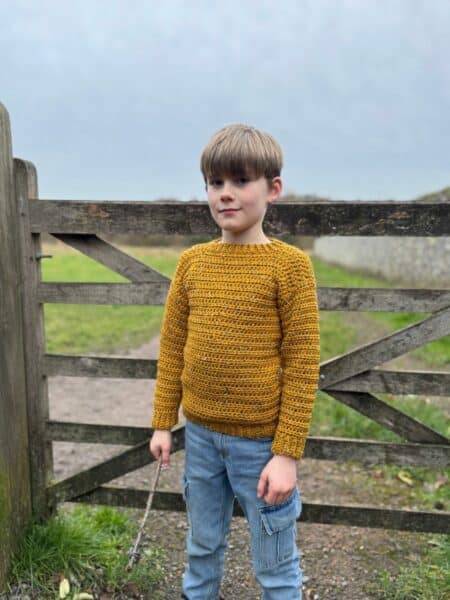 A child in the easiest-to-crochet mustard sweater and jeans holds a stick, standing in front of a wooden fence on a grassy path.