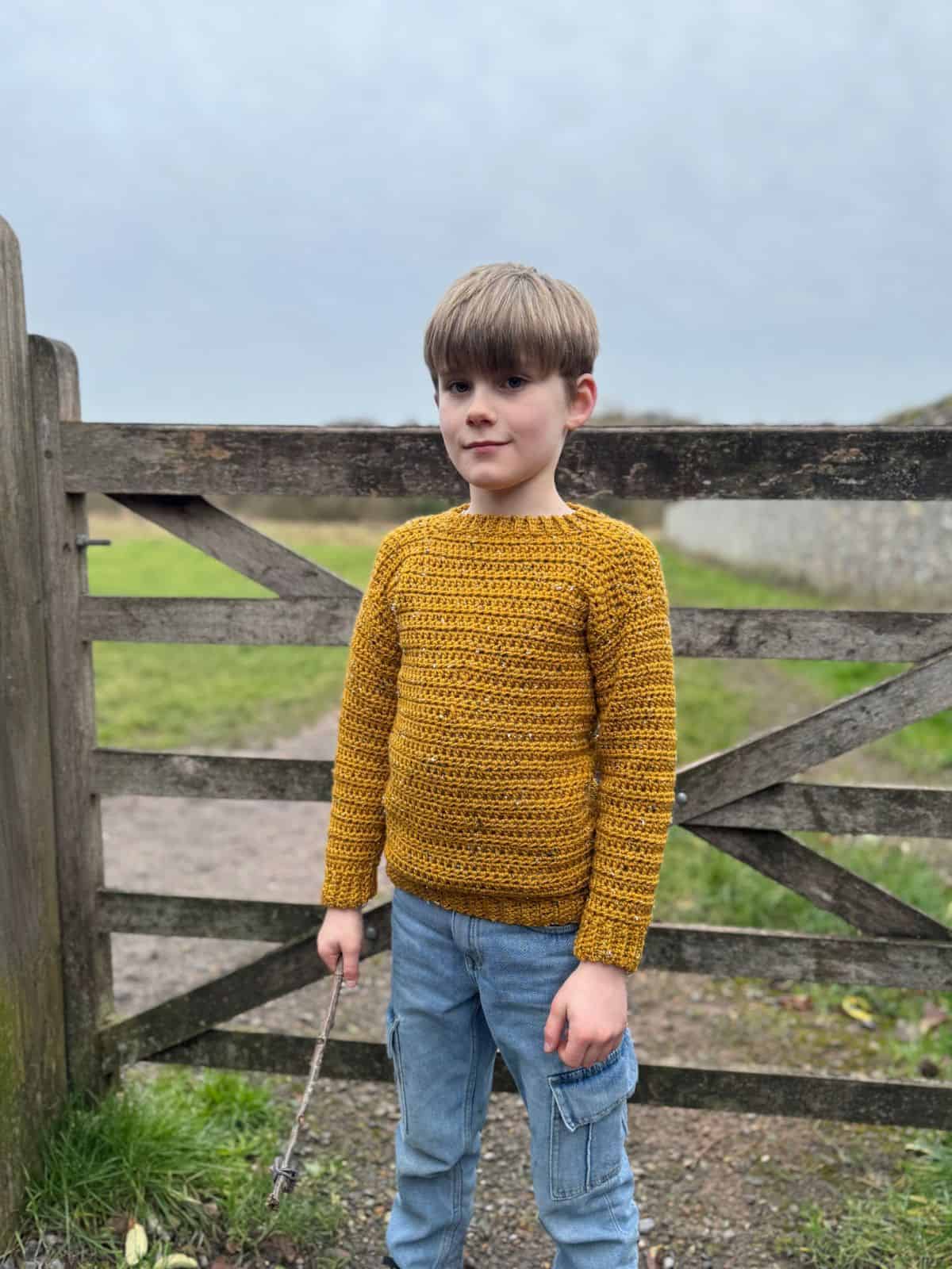 A child in the easiest-to-crochet mustard sweater and jeans holds a stick, standing in front of a wooden fence on a grassy path.