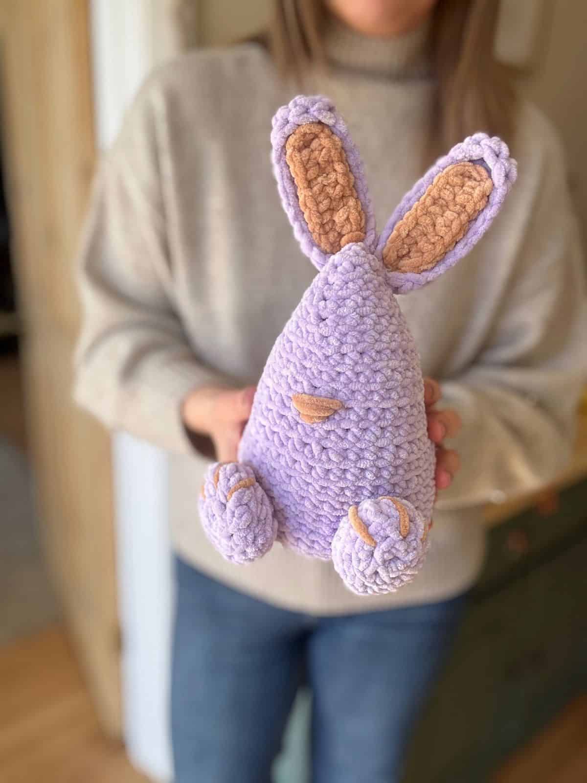 Person holding a crocheted bunny with a lavender body and brown ears, nose, and feet created from a charming crochet bunny pattern.