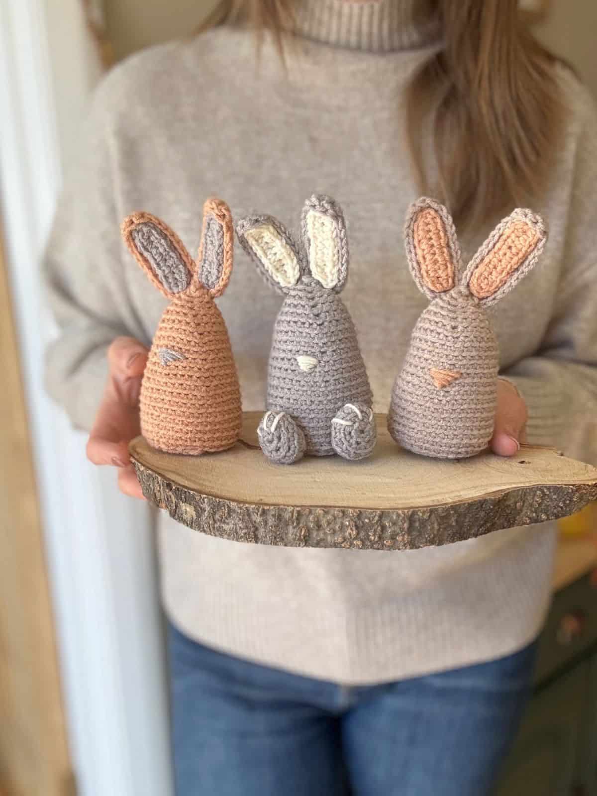 A person displays a wooden slab adorned with three crocheted bunny figures in vibrant colors, each showcasing a unique crochet bunny pattern.