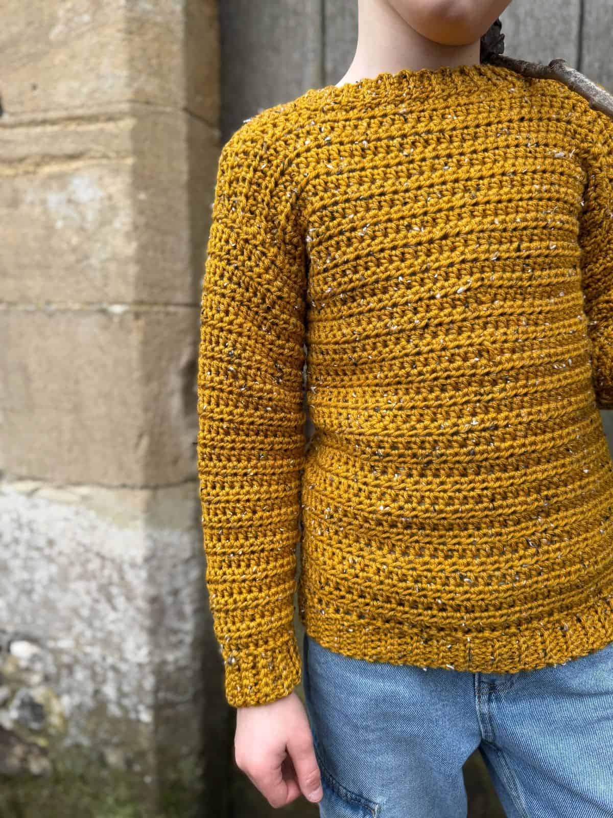 A child stands against a stone wall, wearing a charming yellow sweater reminiscent of the easiest sweater to crochet, paired perfectly with blue jeans.