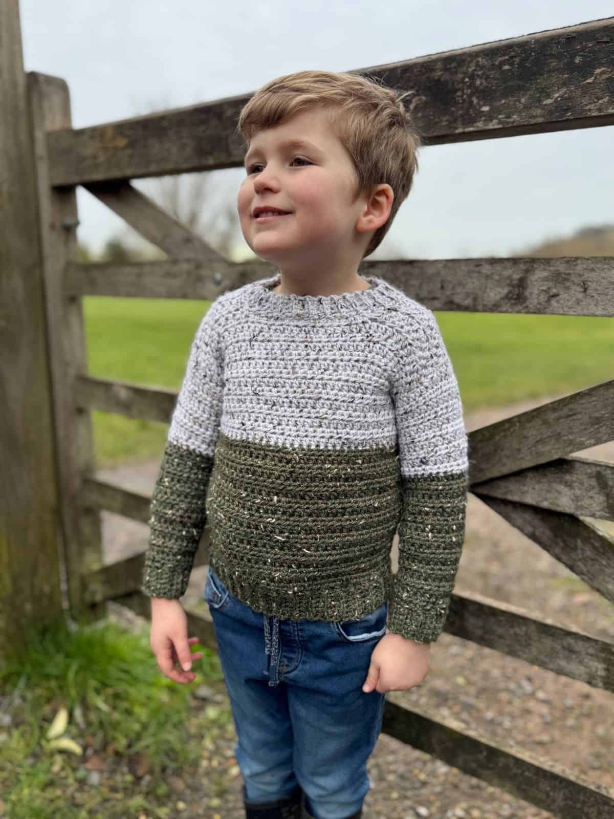 Child in the easiest sweater to crochet, a charming two-toned creation, and jeans stands smiling in front of a wooden gate with a grassy field in the background.