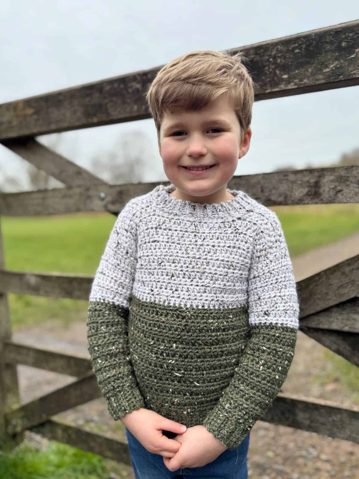 Child in a two-tone crochet sweater stands smiling in front of a wooden fence with grassy fields in the background.