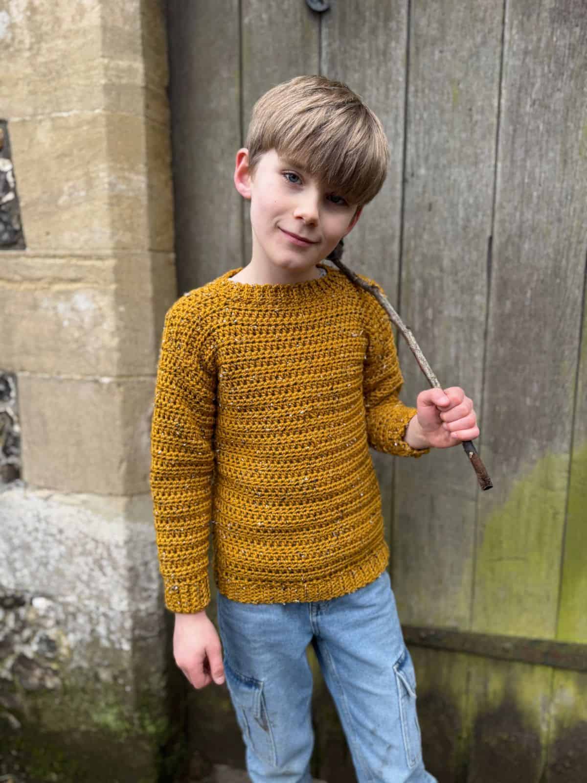 A child, snug in a handmade mustard yellow sweater—the easiest sweater to crochet— and blue jeans, stands in front of a wooden door while clutching a stick.