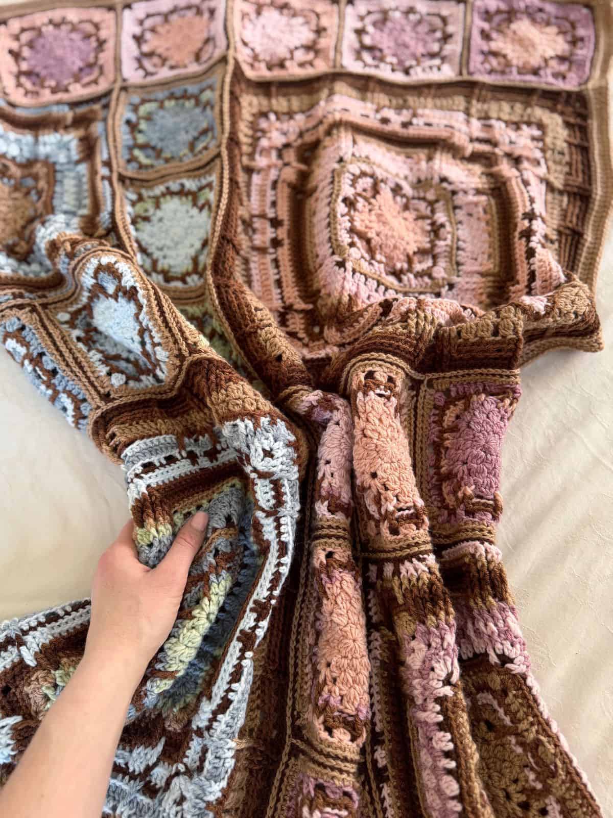 Hand holding a multicolored crochet blanket with square patterns in shades of brown, pink, and blue.