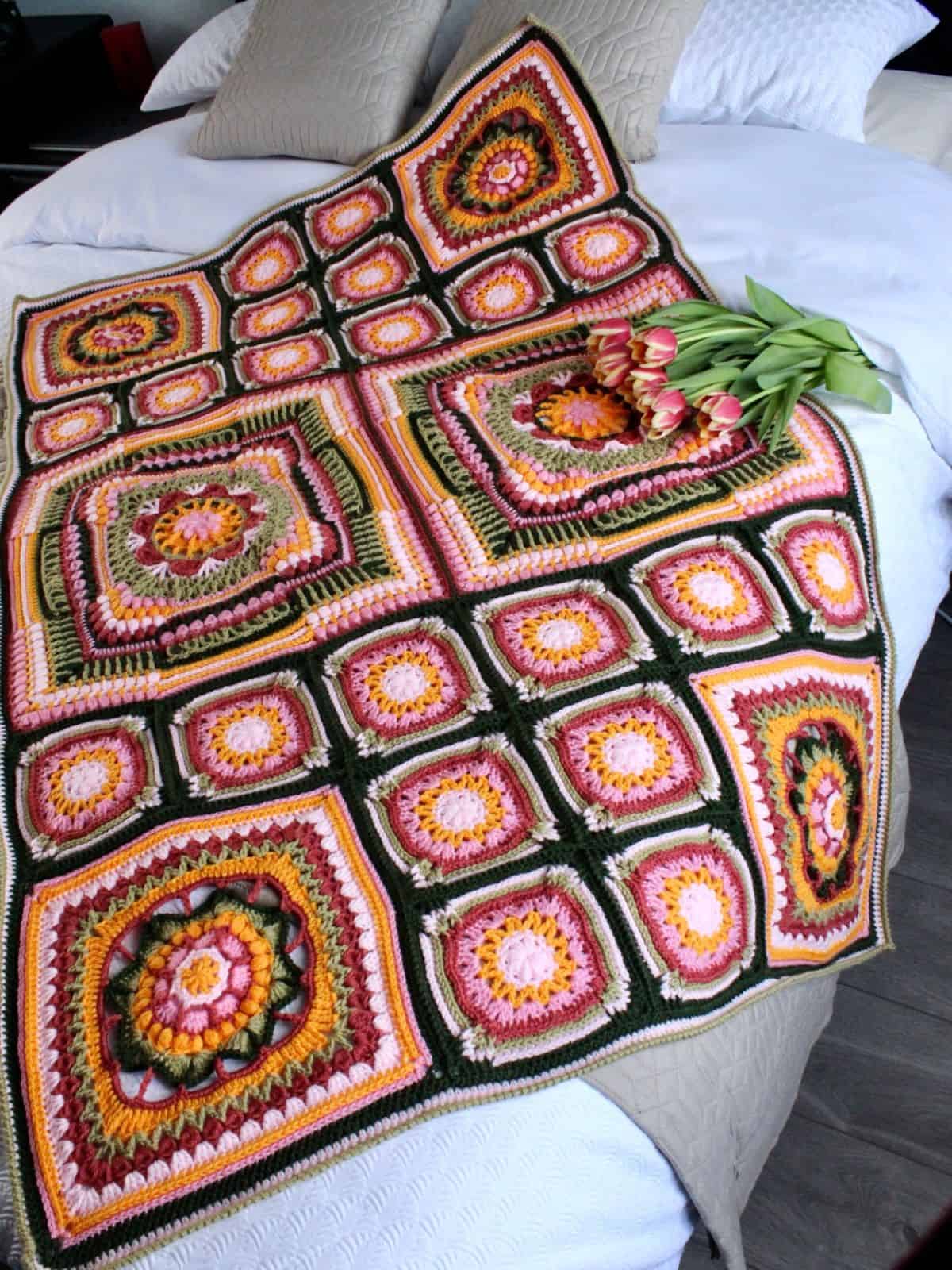 A crocheted blanket with colorful square patterns lies on a bed, accompanied by a small bouquet of tulips.