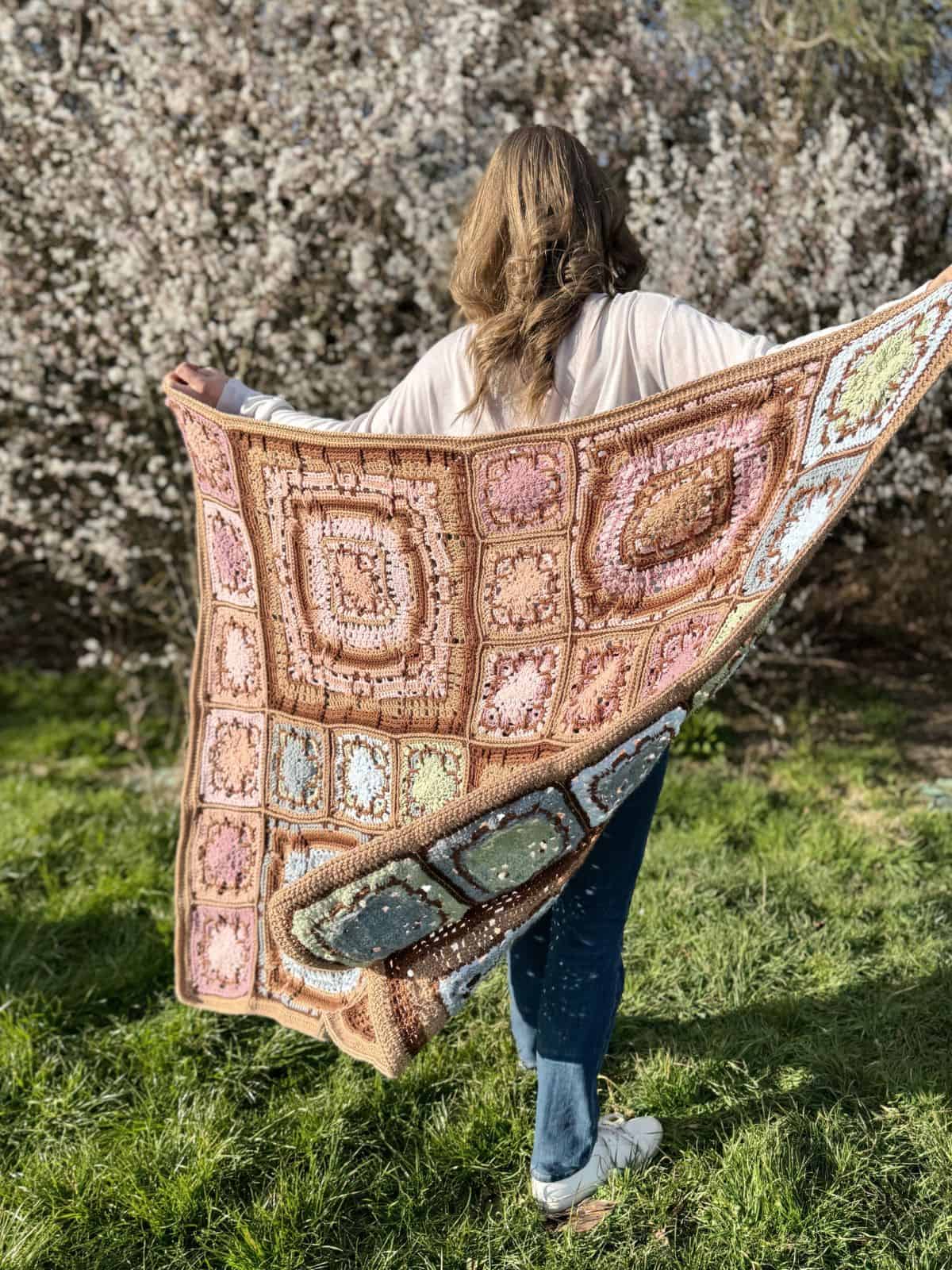 A person stands on grass, holding a colorful crocheted blanket with a block pattern, against a backdrop of blooming trees.