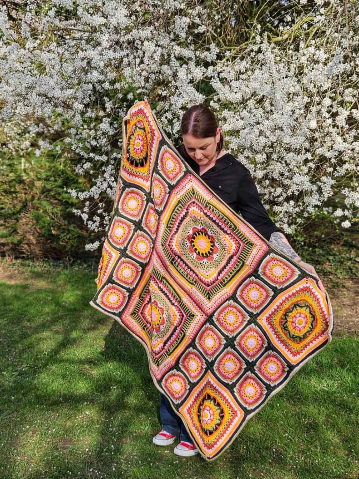 A person stands outdoors holding a colorful crocheted blanket featuring geometric patterns, with a flowering tree in the background.